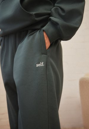 Prim-plan cu o persoană care poartă pantaloni de trening verde închis și hanorac asortat, mâna în buzunar, cu text alb „self.” brodat pe pantaloni.