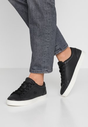 Osoba nosząca czarne skórzane trampki z białymi podeszwami i szare jeansy, jedna stopa lekko uniesiona na jednolitym tle.