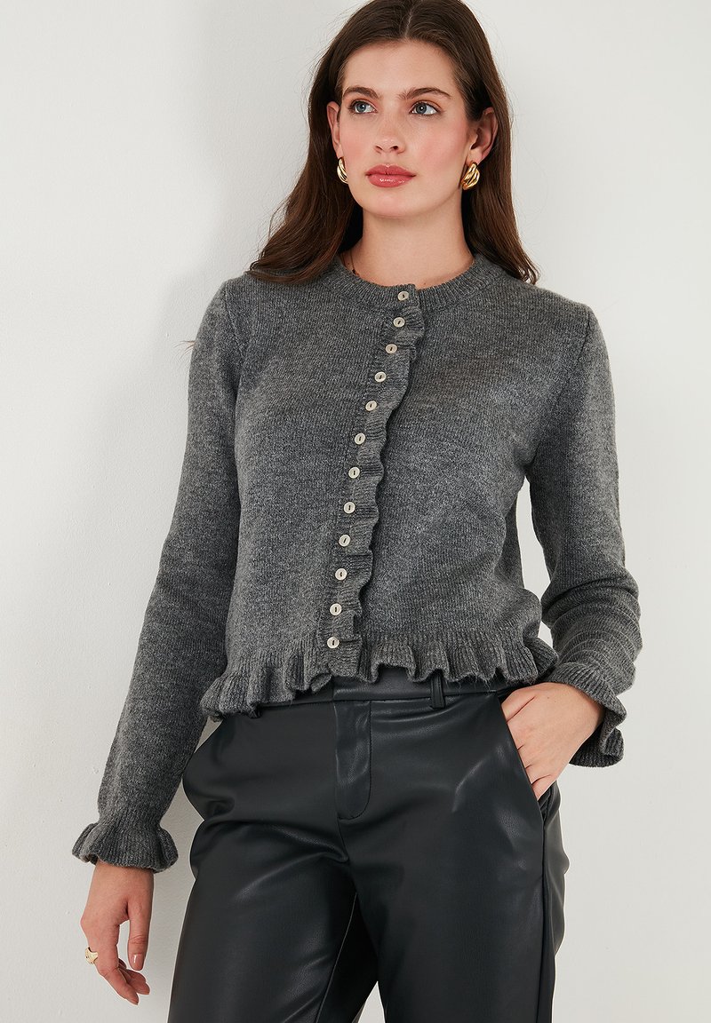 Cardigan grigio a maglia con scollo rotondo, orlo decorativo a balze e chiusura con bottoni. Presenta maniche lunghe e un tessuto testurizzato.