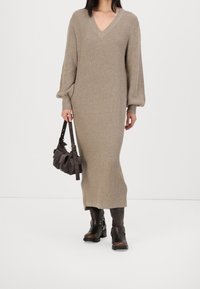 Beige neulottu mekko, jossa on V-pääntie, pitkät puffihihat ja tiukka muoto. Yhdistetty ruskeisiin nilkkureihin ja pieneen tummaan käsilaukkuun.