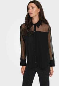 Crna košulja na dugih rukava s prozirnim rukavima, koja ima čvrsti prednji panel i klasični ovratnik. Glatka tkanina s malo labavim krojem.