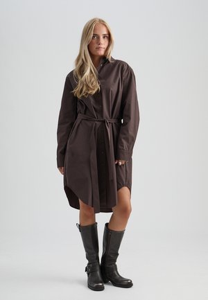 Donna con capelli biondi lunghi che indossa un vestito camicia marrone con bottoni e cintura e stivali neri in pelle al ginocchio, in piedi davanti a uno sfondo semplice.