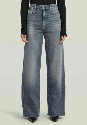 Persona che indossa jeans a vita alta, a gamba larga, di colore blu sbiadito con chiusura a bottoni e cerniera, abbinati a scarpe nere con punta e a una maglia nera a maniche lunghe.