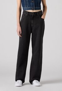 Personne portant un jean noir ample, un gilet en jean court et des baskets blanches, debout devant un fond gris clair uni.