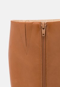 Botas de cuero marrón claro con cremallera lateral y detalle de costura vertical. Textura suave con un brillo sutil.