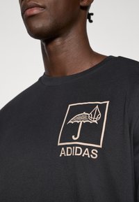 Μαύρο βαμβακερό κοντομάνικο μπλουζάκι με γραφικό λογότυπο που απεικονίζει ομπρέλα και σταγόνα σε ανοιχτό καφέ χρώμα, με το κείμενο "ADIDAS" από κάτω. Στρογγυλή λαιμόκοψη.