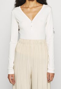 Una persona con lunghi capelli ricci indossa una maglietta bianca a maniche lunghe con scollatura a V e bottoni, abbinata a una gonna plissettata beige.