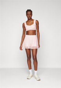Lichtroze sportbh met geplooid detail, gecombineerd met een bijpassende doorzichtige rok. Draag het met witte schoenen en enkelsokken.