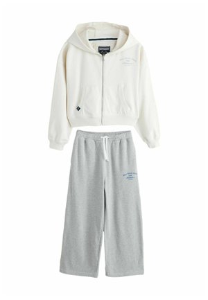 Weißer Reißverschluss-Hoodie mit Kapuze und Kängurutaschen, kombiniert mit grauen weiten Sweatpants mit Logo-Detail und Kordelzugbund.