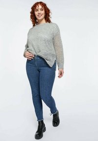Mujer sonriente con cabello rizado rojo, vistiendo un suéter gris de punto suelto, jeans skinny azules con tachuelas y botas negras con cordones caminando hacia adelante.