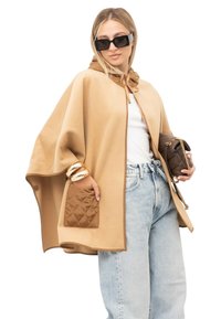 Vrouw draagt beige cape met gewatteerde zak, wit topje, lichtblauwe spijkerbroek, zwarte oversized zonnebril, gouden armbanden en houdt een bruine handtas vast.
