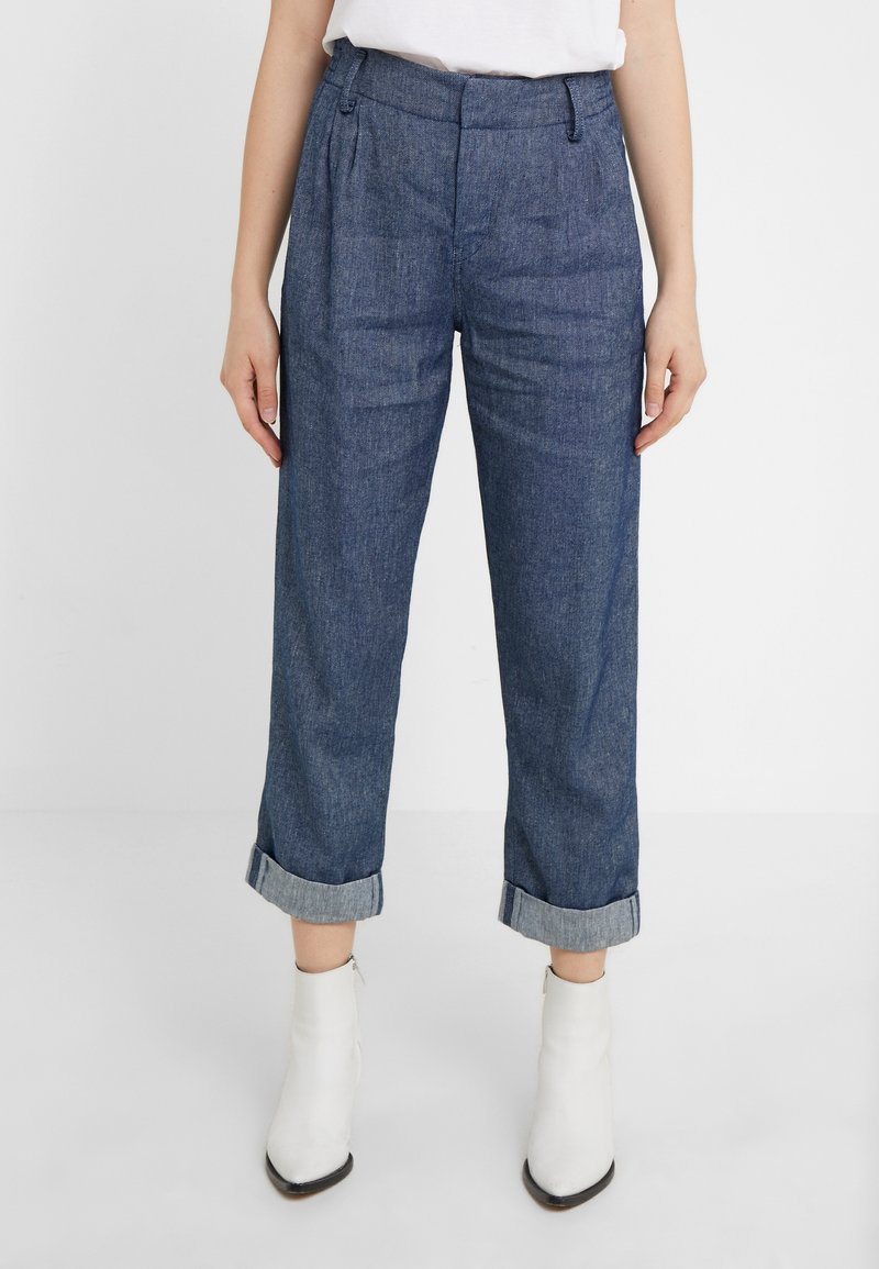 Kvinna som bär högmidjade, uppvikta blå denimbyxor och vita ankelskos, står mot en enfärgad ljus bakgrund.