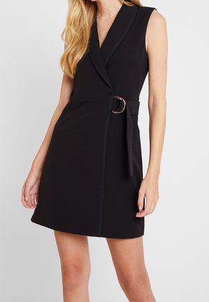 Czarna sukienka bez rękawów typu wrap z kołnierzem z klapą i bocznym paskiem z metalową sprzączką, noszona przez stojącą kobietę z długimi blond włosami.