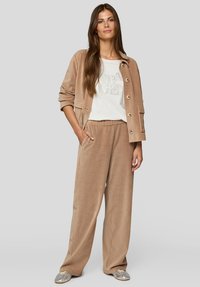 Beige Cordjacke aus Cord mit Knopfverschluss, kombiniert mit passenden weiten Hosen und einem weißen T-Shirt mit silbernem Grafikdruck. Silberne Schuhe.