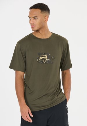 Mann trägt ein olivgrünes T-Shirt mit einem beige-schwarzen Geländewagen-Design, steht mit einer Hand in der Tasche und schaut nach links.