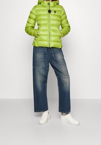 Grøn pufjakke med hætte, der har lynlås og sidelommer, båret over løstsiddende denimjeans og hvide sneakers.