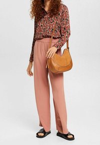 Borsa a spalla in pelle tan con design curvo, camicia a maniche lunghe a fantasia marrone e pantaloni a palazzo rosa chiaro plissettati. Sandali neri scivolati.
