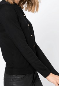 Svart stickad cardigan med rund halsringning, prydd med knappar i guldton längs framsidan och åtsittande ärmar, visad på en modell.