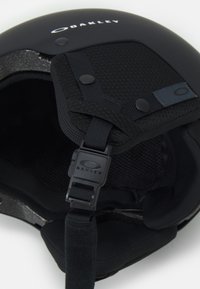 Czarny kask o matowym wykończeniu, wyściełany materiałem, z regulowanym paskiem na brodę i wyraźnym logo Oakley na górze.