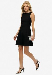 Zwarte mouwloze jurk met een aansluitende bodice en een uitlopende rok, gemaakt van gladde stof. Gecombineerd met zwarte sandalen met bandjes en een metalen clutch.
