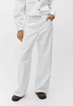 Person iført løse hvide jeans og en hvid knapjakke, med den ene hånd i lommen og brune sko synlige.