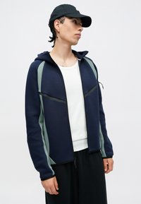 Fato de capuz azul-marinho com detalhes em verde, apresentando um colarinho alto, bolsos com zíper e um tecido texturizado. Usado sobre uma camisa branca.