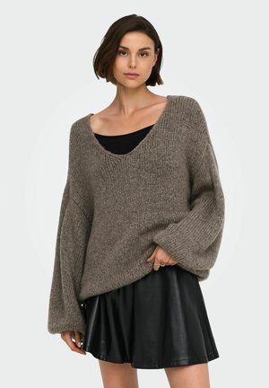 Femme portant un pull en maille gris ample par-dessus un haut noir et une jupe courte en cuir noire, debout devant un fond clair uni.