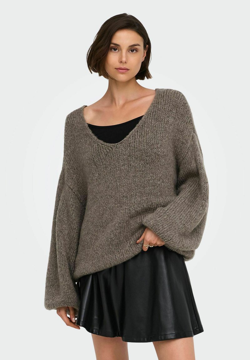 Femme portant un pull en maille gris ample par-dessus un haut noir et une jupe courte en cuir noire, debout devant un fond clair uni.