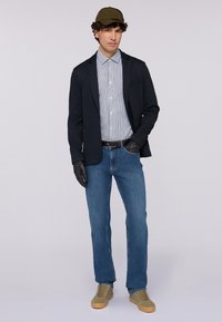 Blazer blu da uomo sopra una camicia a righe, jeans blu e berretto verde oliva. Guanti texture e sneakers marroni chiari completano il look.