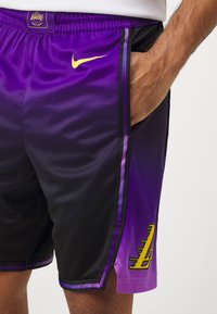 Osoba nosząca czarne i fioletowe spodenki do koszykówki Los Angeles Lakers z żółtym logo Nike i ręką w kieszeni.