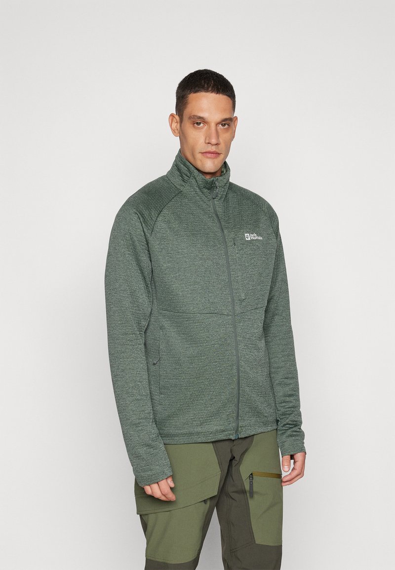 Žalias zip-up džemperis, pagamintas iš textured audinio, su aukštu apykakle, šoninėmis kišenėmis ir logotipu ant krūtinės. Derinamas su chaki spalvos kelnėmis.
