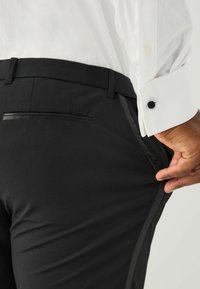 Schwarze Hose mit glatter Textur, flachem Bund und seitlichen Streifen. Eine Gesäßtasche mit schmalem schwarzem Rand ist sichtbar.