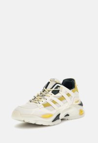 Sneakers chunky in mesh bianco e pelle, con dettagli gialli e neri. Presentano lacci distintivi e una suola ammortizzata. Logo esposto.
