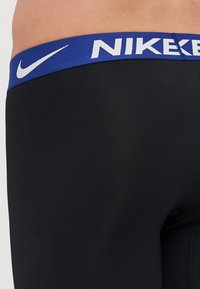 Fekete sportos rövidnadrág kék derékszíjjal, amelyen fehér "NIKE" felirat található. Az anyag sima és testhezálló kialakítású.