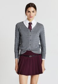 Donna che indossa un cardigan grigio a trecce sopra una camicia bianca con colletto, una cravatta bordeaux e una gonna plissettata bordeaux con una cintura nera.