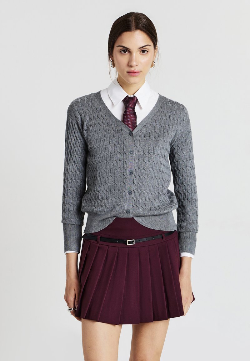 Donna che indossa un cardigan grigio a trecce sopra una camicia bianca con colletto, una cravatta bordeaux e una gonna plissettata bordeaux con una cintura nera.