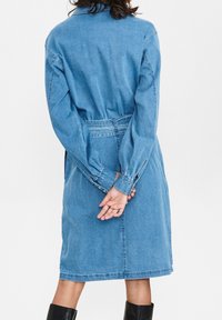Denimklänning i ljusblått, med krage, långa ärmar, midjehögt bälte och knä-lång fåll. Slät textur med knappdetaljer.