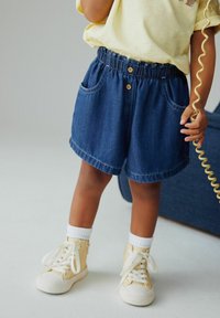 Rugalmas derékpántos farmer rövidnadrág, két oldalzsebbel, világoskék színben, felhajtott szegéllyel; krémszínű sportcipővel és fehér zoknival viselve.