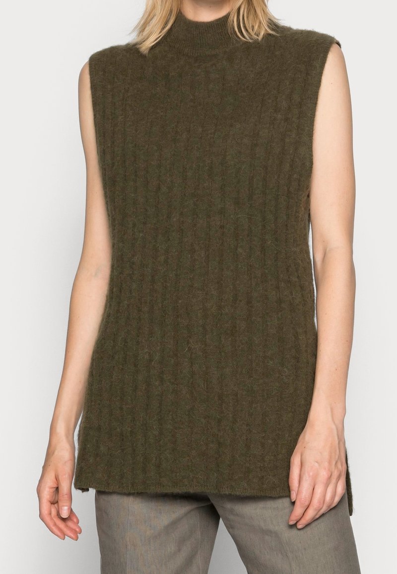 Pull sans manches côtelé vert olive foncé avec un col montant porté avec un pantalon gris, montrant le haut du corps et les bras.