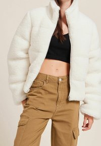 Chaqueta acolchada blanca con forro de vellón, cuello alto y cremallera frontal; combinada con pantalones cargo marrón claro con bolsillos laterales.