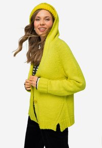 Světlý žlutý pletený cardigan s kapucí, se zapínáním na knoflíky a žebrovanými manžetami. Pod ním je vidět pruhovaná černobílá košile.