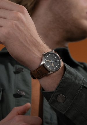 Montre avec un bracelet en cuir marron, un boîtier en acier inoxydable, un cadran gris, des index en argent, et un logo embossé sur le bracelet, portée au poignet.