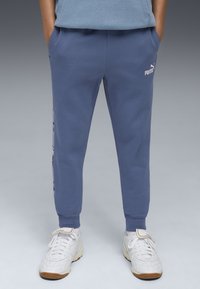 Pantalones de chándal de algodón azul con puños de canalé, bolsillos laterales y detalle de logo en blanco. Combinados con zapatillas blancas con suela marrón.