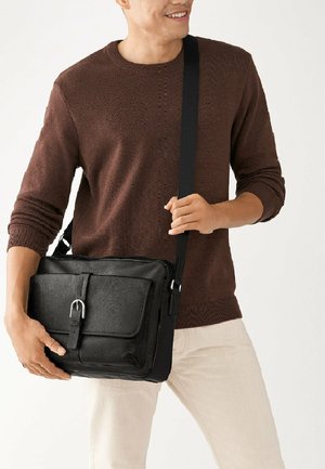 Schwarze Leder-Umhängetasche mit strukturierter Oberfläche, Schnallenverschluss, verstellbarem Tragegurt, gehalten von einer Person, die einen braunen Strickpullover trägt.