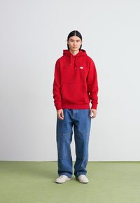 Hanorac roșu cu glugă, prevăzut cu un buzunar frontal și șnururi, asortat cu blugi largi de culoare albastru deschis și adidași albi, pe o suprafață verde.