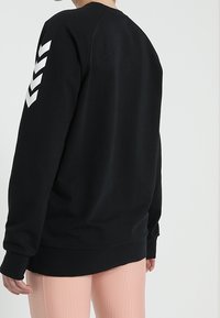 Sweatshirt noir à manches raglan, avec des bandes en chevron blanches sur la manche gauche, poignets et ourlet côtelés, fabriqué en tissu doux.