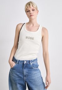 Crème tanktop met geribbelde textuur met daarop "BOSS" in strass, gecombineerd met lichtblauwe high-waisted denim jeans.