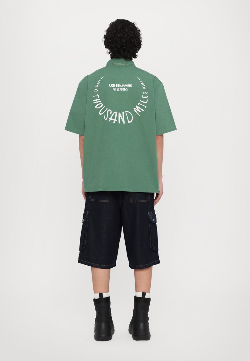 Zelena majica kratkih rukava s belim natpisom na leđima, uparena s tamnoplavi denimski cargo šortsevi i crnim visokim čizmama. Glatka pamučna tekstura.