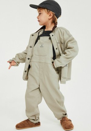 Jong kind draagt beige tuinbroek, bijpassend jasje, zwart shirt, zwarte pet en bruine schoenen, staat met één hand in de zak en wijst met de andere.