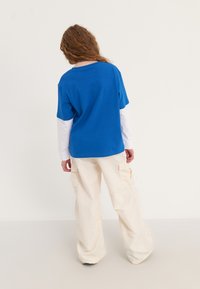 Loose blue t-shirt over a white long-sleeve shirt, paired with off-white cargo pants. Curly hair falls over shoulders, minimal design details.
